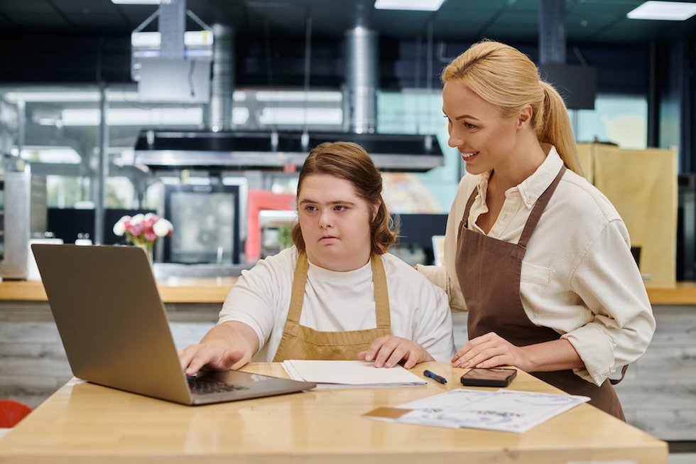 Administrador alegre sonriendo junto a una empleada con síndrome de Down que trabaja con un ordenador portátil en una cafetería.